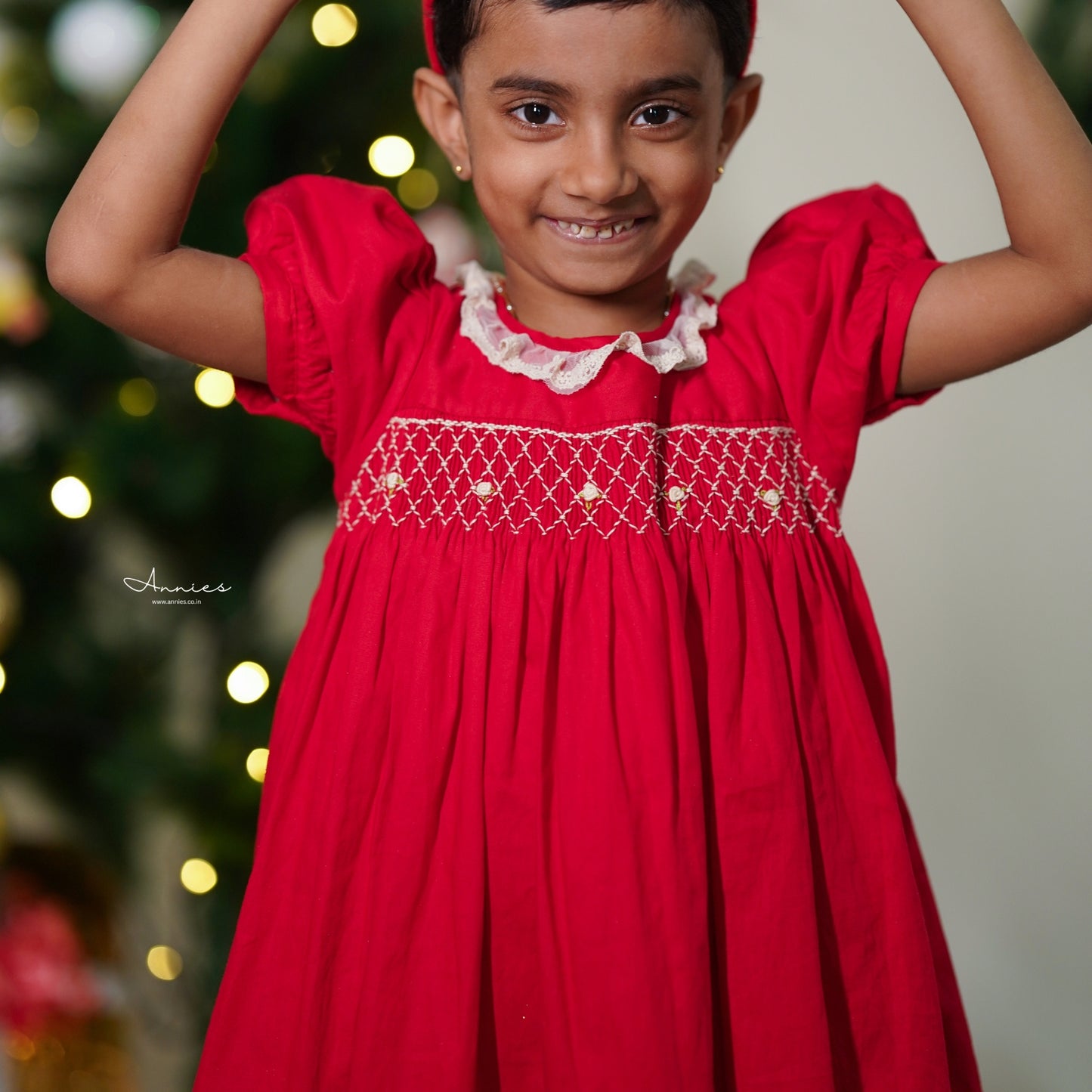 Young girl wearing a red dress with white lace details, standing against a blurred festive background.