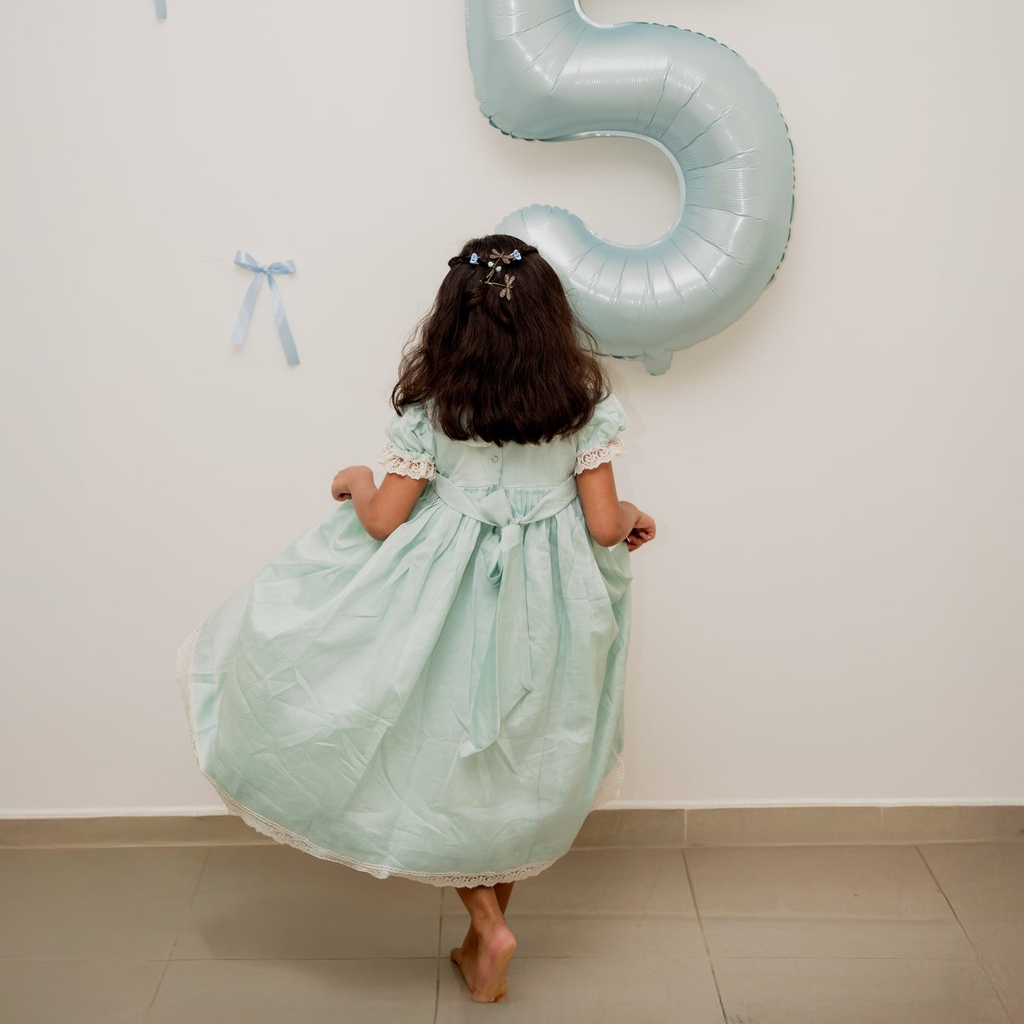 Child in a light green dress holding a large blue number 5 balloon against a white wall.