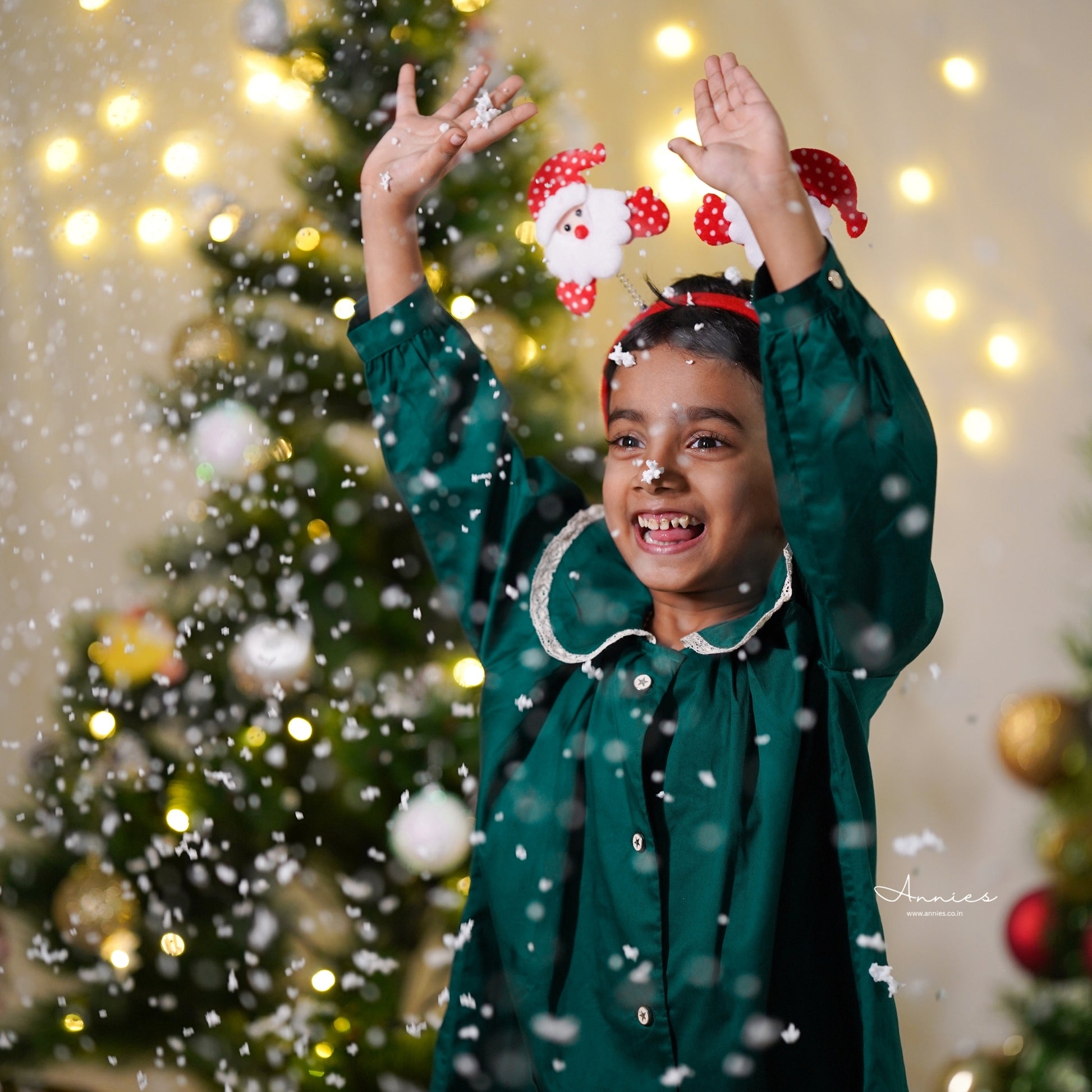 Child in a green outfit with festive headband in front of a decorated Christmas tree with lights.