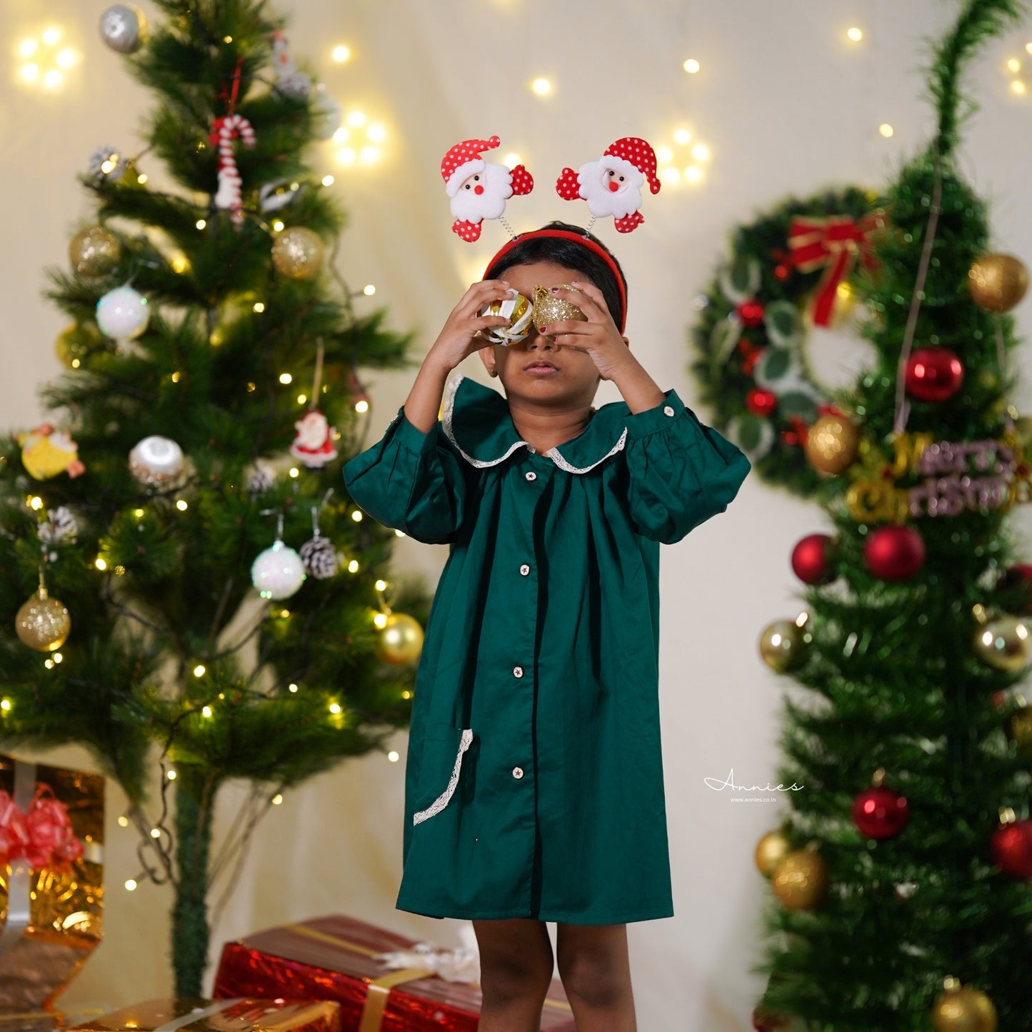 Child in a green dress with festive headband in front of Christmas trees and decorations.
