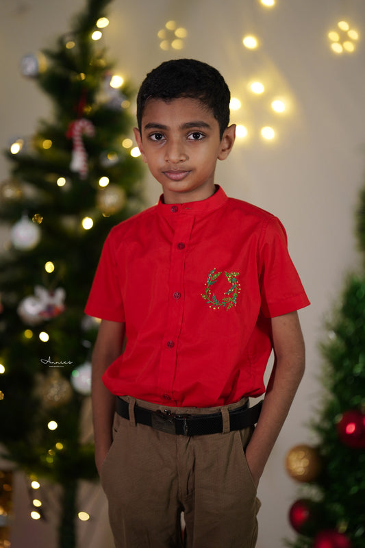 Young boy wearing a red shirt with a Christmas design in front of a decorated tree.