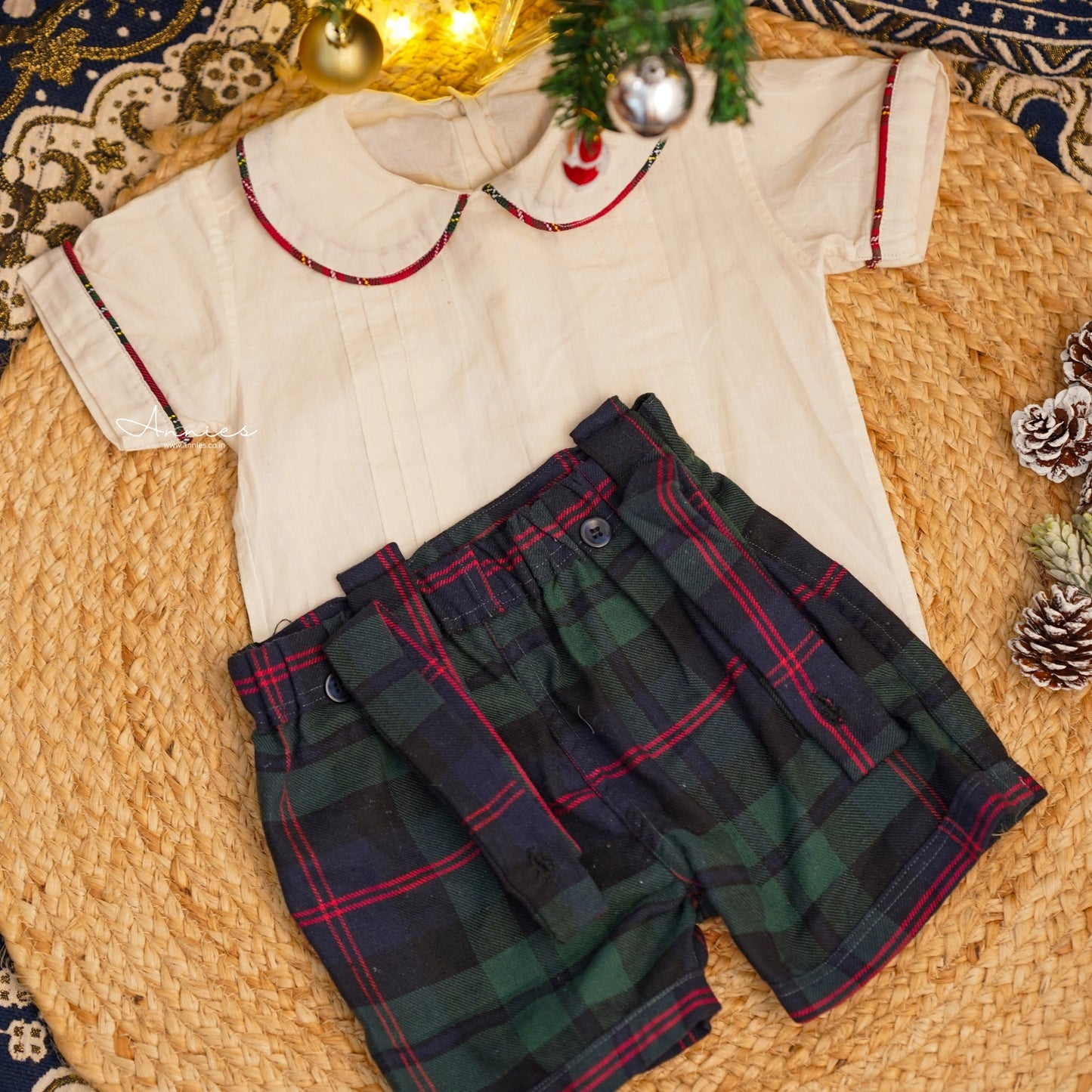 Children's outfit with white shirt and plaid shorts on a textured surface with Christmas decorations.