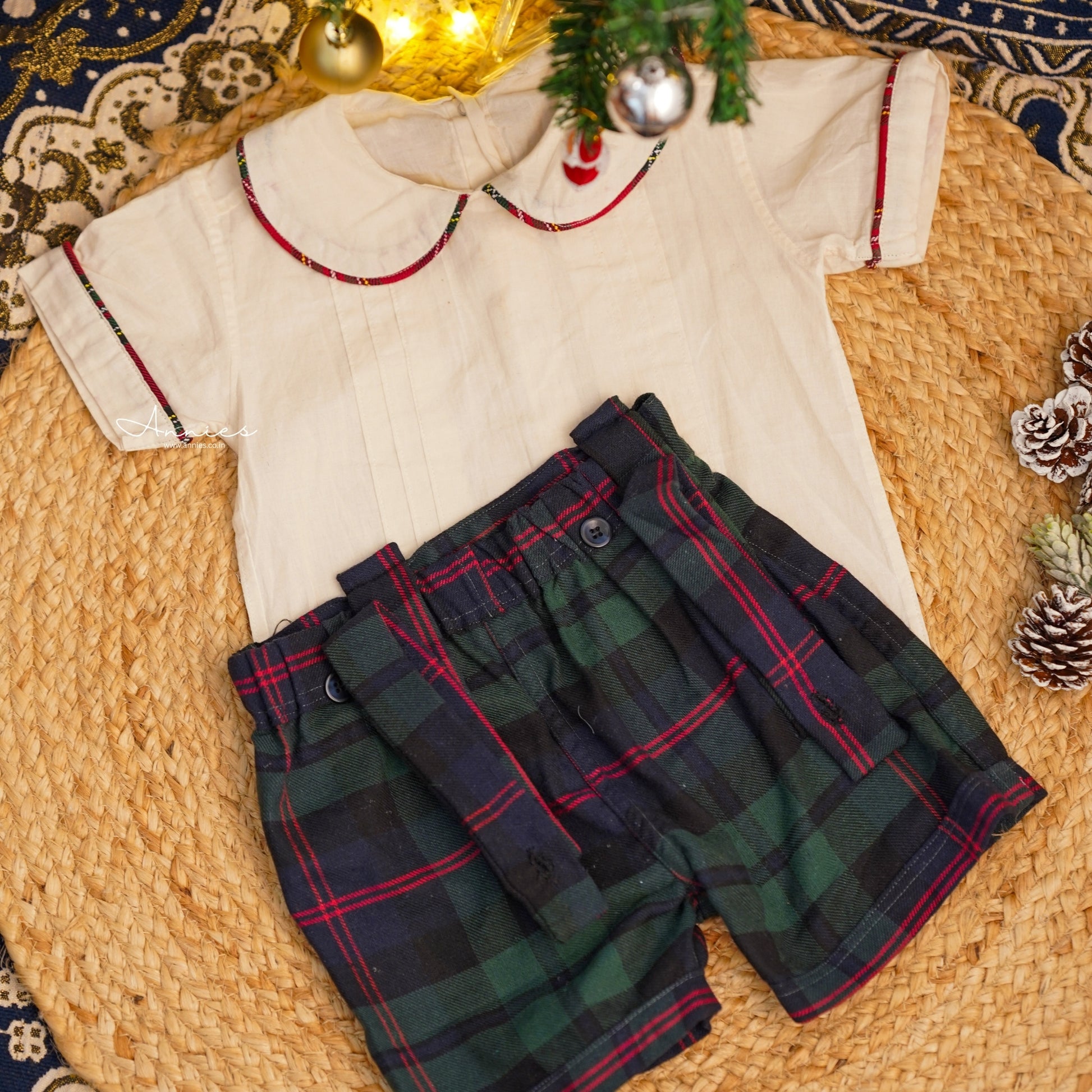 Children's outfit with white shirt and plaid shorts on a textured surface with Christmas decorations.