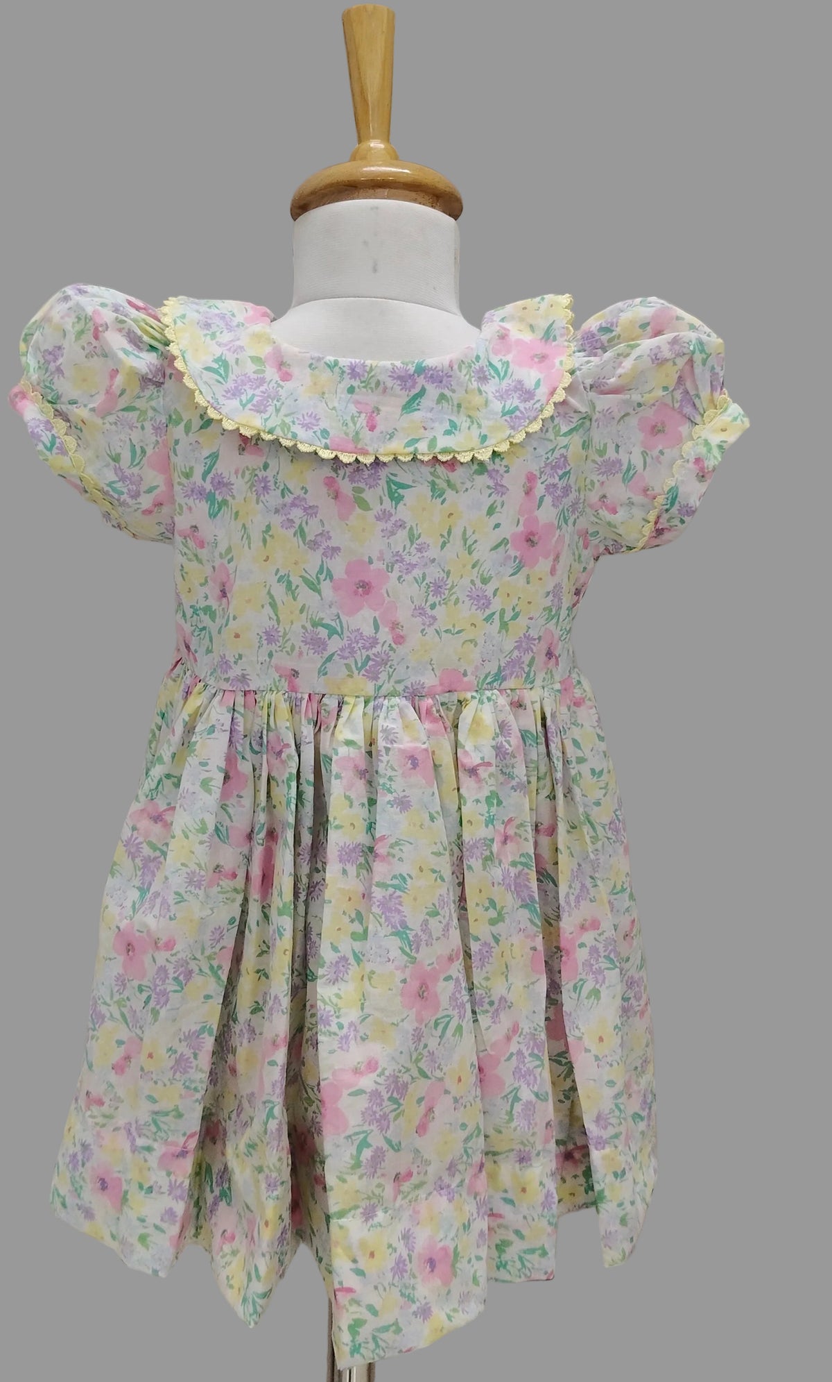 A toddler girl's short-sleeve dress with a pastel floral print, displayed on a white dress form. The dress features a ruffled yellow-trimmed neckline, puffed sleeves, and a gathered skirt, shown against a background of beige horizontal slats.