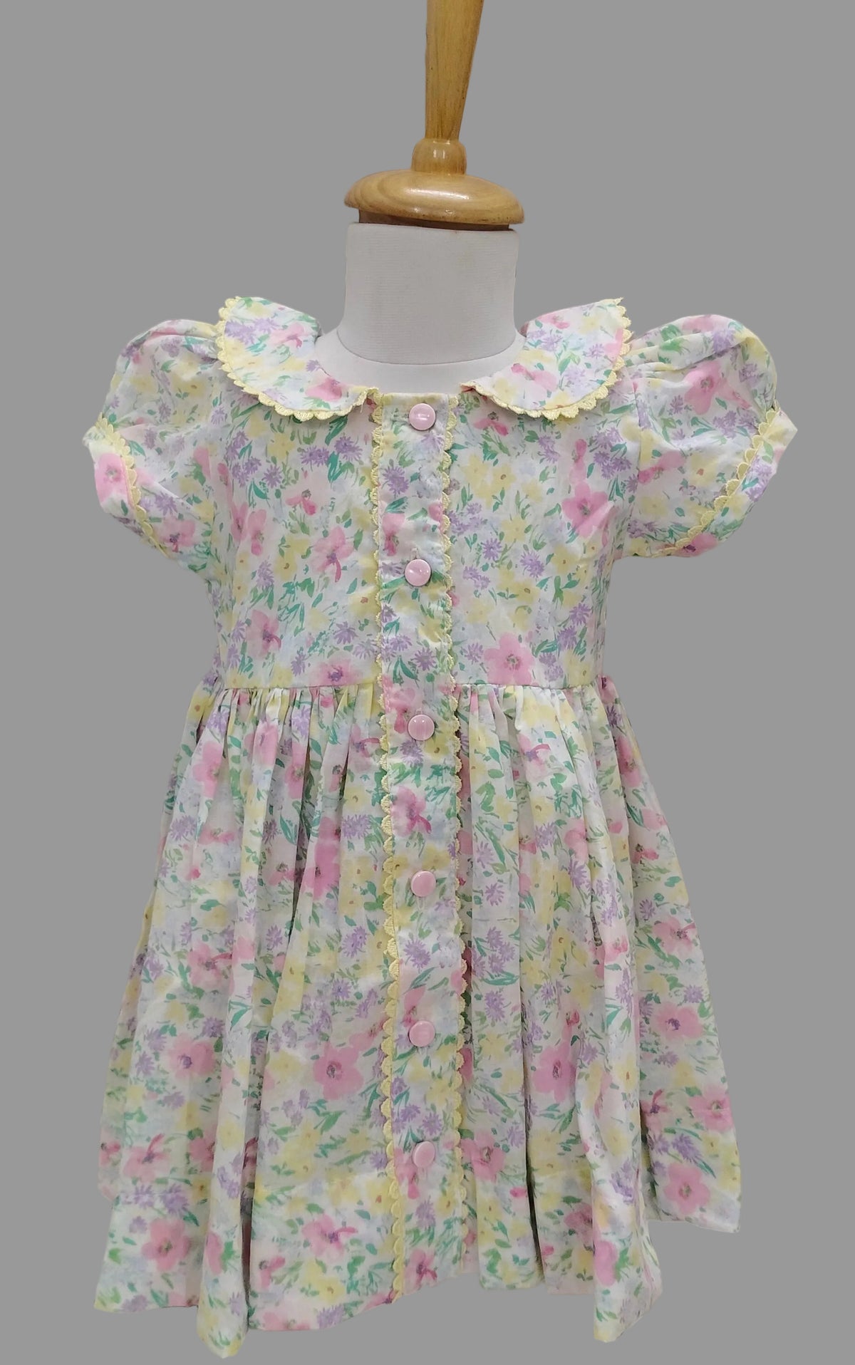 A toddler girl's short-sleeve dress with a pastel floral print, displayed on a mannequin dress form against a beige horizontal slat background. The dress features a ruffled yellow collar, buttoned front, and a gathered skirt.