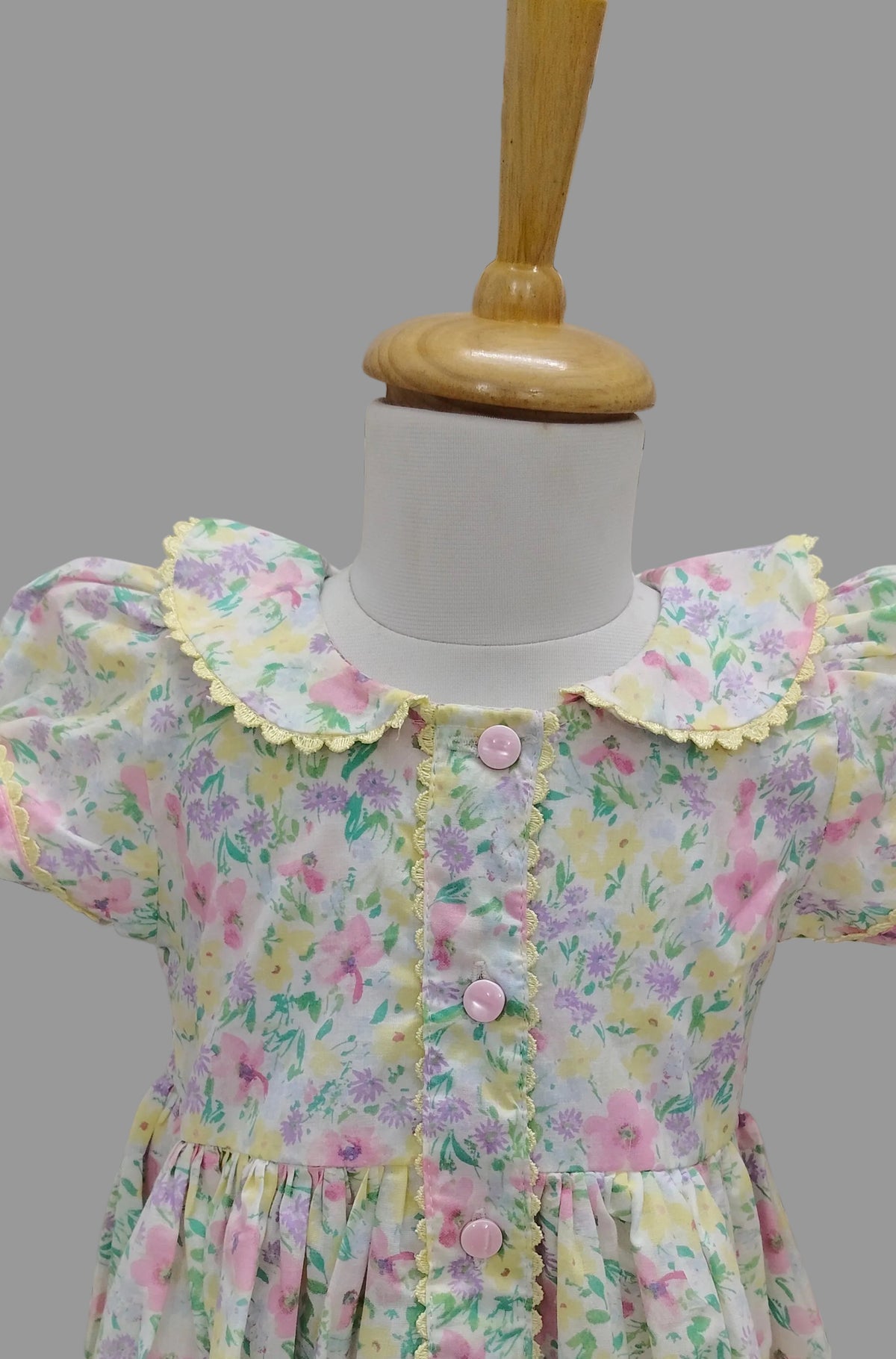 A short-sleeve girl's dress with a pastel floral print, displayed on a white mannequin torso with a wooden neck cap. The dress features a yellow ruffled Peter Pan collar, pink buttons, and a matching ruffled waistband.