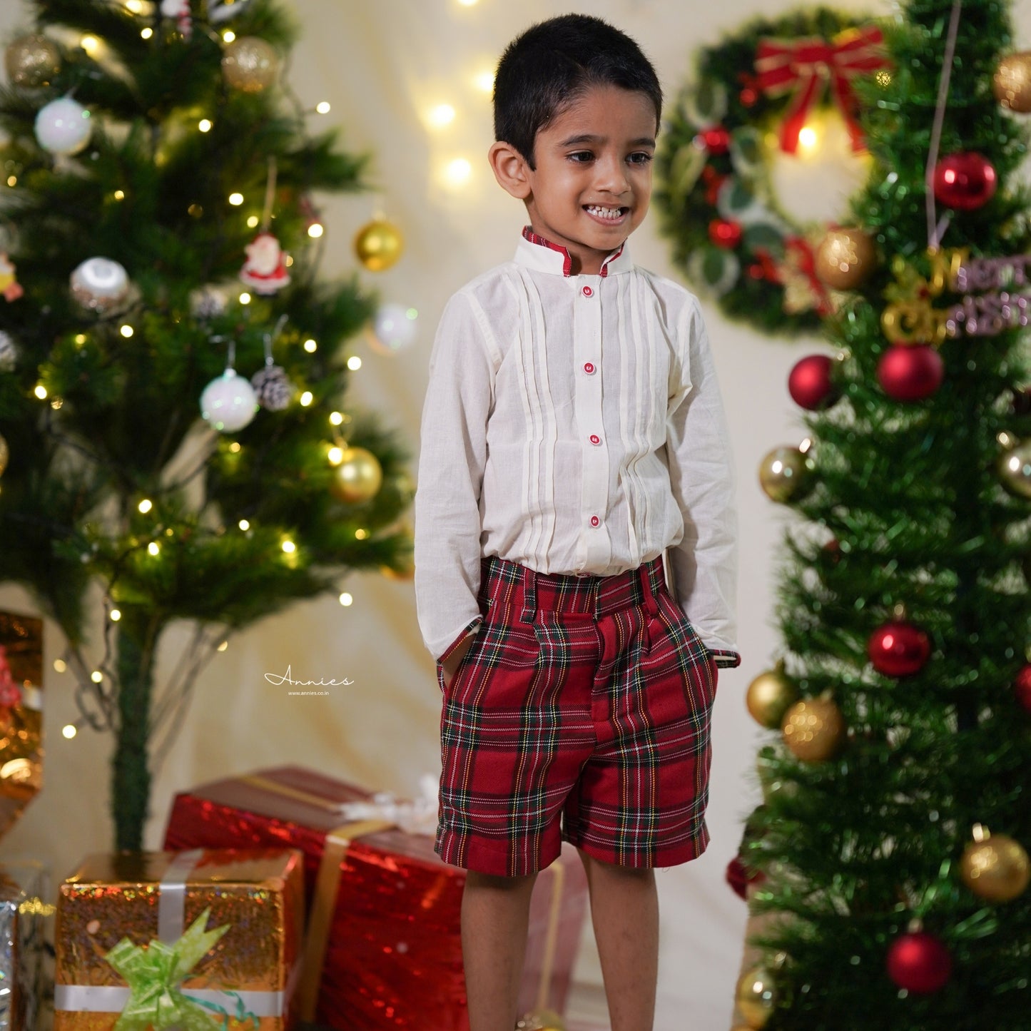 Child in festive attire standing between two decorated Christmas trees with lights and presents.