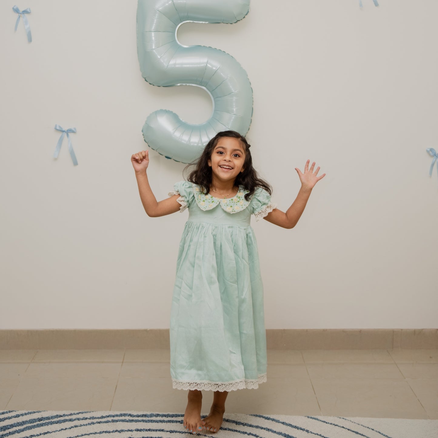 Young girl in a light green dress standing next to a large number five balloon indoors.