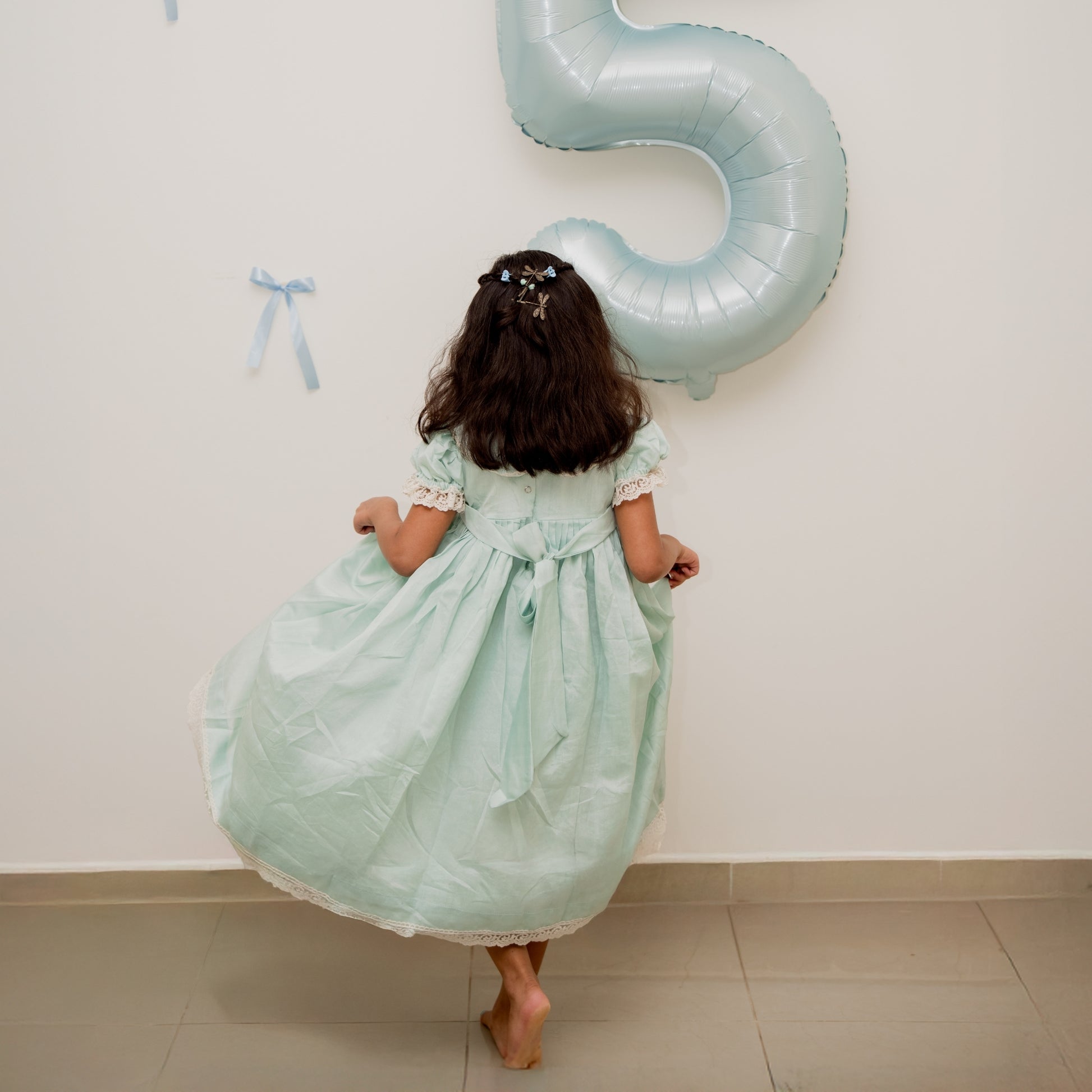 Child in a light green dress holding a large blue number 5 balloon against a white wall.